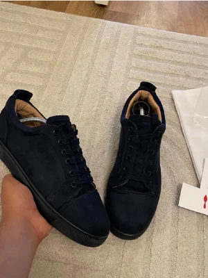 Mörkblå sneakers från Christian Louboutin - Säljer ett par mörkblå sneakers från Christian Louboutin i mjuk mocka med rund tå och platt sula. Skorna har snörning och diskret broderad logga på hälen. Insidan är beige och detaljerna är stilrena. Perfekt för dig som gillar lyxiga och snygga sneakers. Obs ett litet hål finns vid hälen 