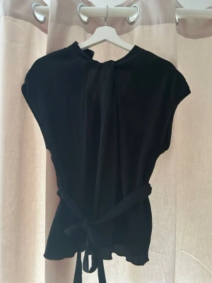 Svart omlottopp med knytband - Snygg svart omlottopp med knytband i midjan och lätt ribbad struktur. Toppen har korta ärmar och en avslappnad passform som ger en stilren look. Perfekt att styla med jeans eller kjol för en trendig outfit.