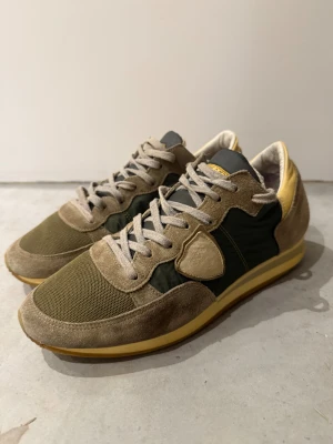 Philippe Model  - Säljer ett par snygga Philippe Model sneakers i olivgrönt och beige med detaljer i gult. Ovandelen är i mocka och nylon, med mesh framtill och klassisk snörning. Skorna har en platt gummisula och rund tå. Perfekta för dig som gillar retrovibbar och streetstyle. Storlek 40 men passar 41 möjligtvis 41,5