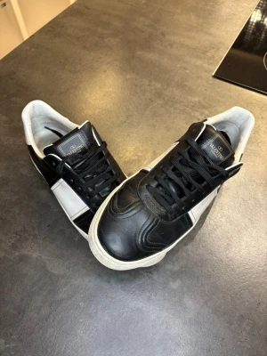 Valentino Flycrew - Säljer ett par snygga Valentino sneakers i svart och vitt skinn. Skorna har klassisk snörning, rund tå och coola nitar på hälen. Tidlös design med kontrasterande paneler och logga på plösen. Perfekta för dig som vill ha en lyxig touch till din outfit.