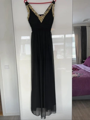 Svart långklänning med guldstenar - Säljer en svart långklänning med v-ringad front och breda axelband. Klänningen har glittriga guldpaljetter längs urringningen och axelbanden, samt ett plisserat liv. Kjolen är luftig och faller snyggt. Perfekt för bal eller festkväll!