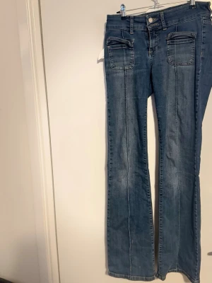 Blå bootcut jeans med fickdetaljer  - Säljer ett par blå jeans med bootcut-modell och markerade sömmar längs benen. Jeansen har två fickor framtill med ”lock” samt klassisk knappgylf. 