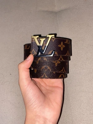 Brun monogram skärp - Säljer ett klassiskt skärp i brunt med ikoniskt monogrammönster och stor logga i guld och svart metall. Bältet är gjort i skinn och har beige insida. Perfekt accessoar för att lyfta vilken outfit som helst.