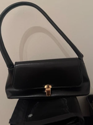 Svart axelväska i skinn - Snygg svart axelväska i skinn med stilren, avlång form och guldfärgat spänne framtill. Väskan har ett klassiskt handtag och minimalistisk design som passar till många olika looks. Perfekt för dig som gillar en clean och tidlös stil.