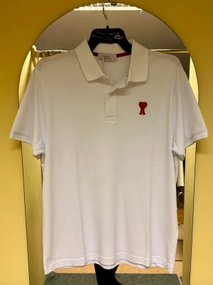 Vit pikétröja från Ami med rött hjärta - Klassisk vit pikétröja från Ami med krage och knappar framtill. På bröstet sitter Ami:s ikoniska röda hjärtemblem. Tröjan har korta ärmar och är gjord i mjuk bomull som känns skön mot huden. Perfekt för en clean och stilren look.