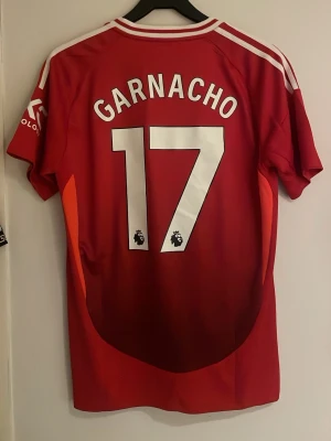 Röd Manchester United matchtröja - Säljer en röd Manchester United matchtröja med Garnacho och nummer 17 på ryggen. Tröjan har vita detaljer på axlarna, klubbmärke på bröstet och Snapdragon som sponsortryck. Tillverkad i lätt och ventilerande polyester, perfekt för fotbollsplanen.
