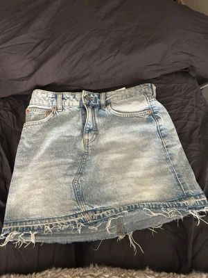 Ljusblå jeanskjol med fransig kant - Säljer en ljusblå jeanskjol i klassisk modell med fickor och som är fransig med rå kant nedtill. Kjolen har bälteshällor och knappstängning framtill. Perfekt för dig som gillar en avslappnad och trendig denimlook.