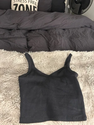 Svart ribbad linnetopp - Säljer en svart ribbad linnetopp med smala axelband och v-ringad hals. Toppen är croppad och har en enkel, stilren design som passar perfekt till jeans eller kjol. Materialet är mjukt och stretchigt för en bekväm passform.