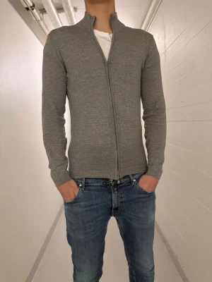 Grå kofta med dragkedja - Säljer en stilren grå kofta med hel dragkedja och hög krage. Tröjan är långärmad och har en slim passform som sitter snyggt på kroppen. Perfekt att slänga på sig över en t-shirt för en clean look.