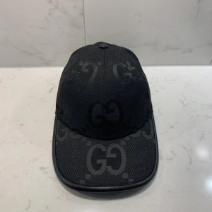 Svart keps från Gucci med GG-mönster - Säljer en svart keps från Gucci med ikoniskt GG-mönster ton-i-ton över hela kepsen. Kepsen har böjd skärm och justerbar rem baktill. Tillverkad i ett slitstarkt tygmaterial som ger en exklusiv känsla.