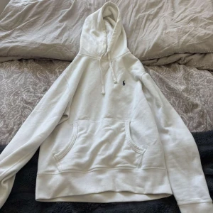 Vit hoodie från Polo Ralph Lauren - Säljer en stilren vit hoodie från Polo Ralph Lauren med klassisk broderad logga på bröstet. Tröjan har huva med snörning, känguruficka och är tillverkad i mjuk bomull. Perfekt för en chill och clean look.