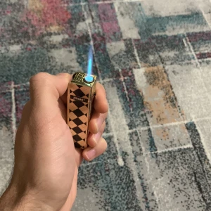 Jeddah Elektronisk Tändare - Stilren elektronisk tändare från Jeddah med diamantmönster. Ger en kraftfull blå låga och är enkel att använda. Laddningsbar via USB. Mycket gott skick, inga synliga repor eller skador. Perfekt för dagligt bruk eller som present.