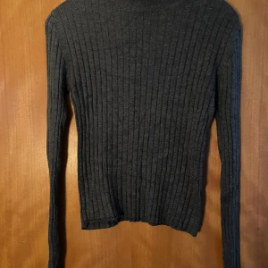 Mörkgrå ribbad polotröja - Säljer en mörkgrå ribbad polotröja med lång ärm. Tröjan är figurnära och har en klassisk hög hals. Perfekt att styla med jeans eller kjol för en clean look.