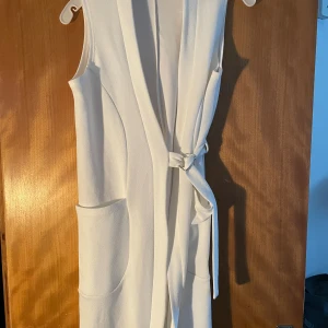 Vit ärmlös omlottklänning med knyt - Snygg vit omlottklänning med knyt i midjan och stora fickor framtill. Klänningen är ärmlös och har en stilren, modern look som passar perfekt för varma dagar. Materialet känns mjukt och har en lätt struktur. Enkel att matcha med accessoarer för en personlig stil.