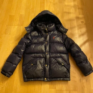 Svart pufferjacka från Polo Ralph Lauren - Mörkblå Ralph Lauren pufferjacka storlek M 152 Har ett litet hål i ryggen (se sista bilden). 