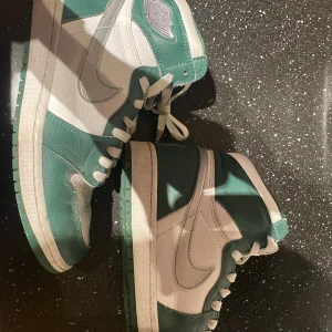 Nike Air Jordan 1 gröna och vita - Säljer ett par Nike Air Jordan 1 i en snygg grön och vit färgkombination. Skorna har klassisk high-top design, snörning och är tillverkade i skinn med vit swoosh på sidorna. Perfekta för dig som vill sticka ut med en ikonisk sneaker.