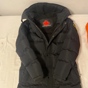 Svart pufferjacka från Zavetti Canada - Snygg svart pufferjacka från Zavetti Canada med stor huva och broderad patch på ärmen. Jackan har dragkedja och knappar framtill, samt ett fluffigt foder som håller dig varm. Perfekt för kalla dagar och har en cool look med tydliga quiltade sektioner.