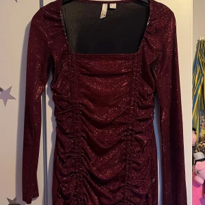 Glittrig vinröd klänning med rynk - Supersnygg vinröd glittrig klänning från Body Flirt med lång ärm och fyrkantig urringning. Klänningen är kort och har rynkade detaljer framtill som ger en cool och trendig look. Perfekt för dig som vill sticka ut med lite extra glitter. En perfekt julklänning🎄