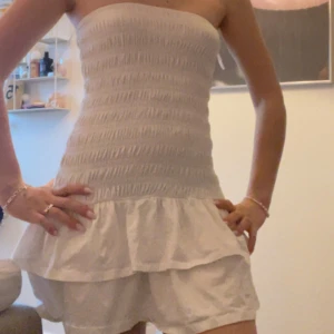 Vit axelbandslös kortklänning - Supersöt vit axelbandslös klänning med smock upptill och volang nedtill. Perfekt för varma dagar och har en luftig känsla. Klänningen är kort och har en feminin siluett som framhäver axlarna. Materialet känns lätt och bekvämt mot huden.