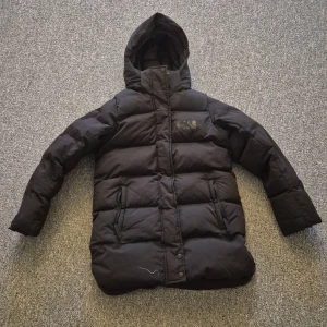 Svart dunjacka från Helly Hansen - Svart dunjacka från Helly Hansen med huva och långa ärmar. Jackan har två sidofickor med dragkedja och tryckknappar framtill. Puffad design med diskret logga på bröstet och bak på nederkanten. Perfekt för kalla dagar och har en clean, sportig look. Nästan som ny.