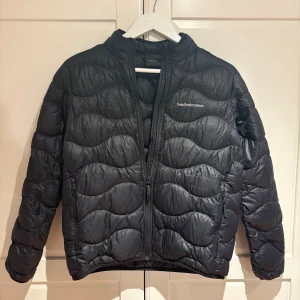 Svart dunjacka från Peak Performance - Svart dunjacka från Peak Performance med vågigt quiltat mönster och diskret logga på bröstet. Jackan har hög krage, två sidofickor med dragkedja och långärmade ärmar. Perfekt för kalla dagar och har en snygg, sportig look.