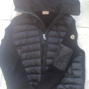 Moncler Cardigan - Snygg svart cardigan från Moncler med stickade ärmar och vadderad kropp. Jackan har huva, dragkedja framtill och en praktisk ficka med dragkedja på ena ärmen. Perfekt för kyliga dagar och riktigt stilren look.