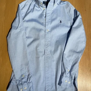 Blå skjorta från Polo Ralph Lauren storlek 16(junior) slim fit.  - Snygg ljusblå skjorta från Polo Ralph Lauren med klassisk krage och knappar framtill. Skjortan har lång ärm med manschett och den ikoniska broderade loggan på bröstet. Tillverkad i mjuk bomull. Använd ett fåtal gånger. 