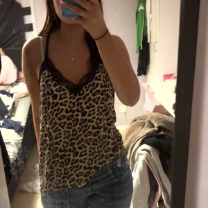 Leopardmönstrad linnetopp med spets - Säljer en snygg leopardmönstrad linnetopp med svarta spetsdetaljer vid halsringningen. Toppen är ärmlös och har tunna axelband, perfekt för dig som gillar djurmönster och vill sticka ut lite extra. Passar till både jeans och kjol!