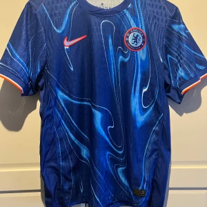 Chelsea Palmer #20 fotbollströja Nike - Säljer en officiell Chelsea FC matchtröja från 24/25 med Palmer och nummer 20 på ryggen. Tröjan är marinblå med unikt abstrakt mönster i ljusblå toner, korta ärmar och orange detaljer vid mudden. Materialet är lätt och andas, perfekt för match eller träning.