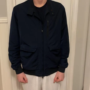 Marinblå vindjacka från J.Lindeberg - Snygg marinblå vindjacka från J.Lindeberg med klassisk krage, dragkedja och tryckknappar. Jackan har två stora fickor framtill, en bröstficka med dragkedja och ribbade muddar vid ärmslut och nederkant. Perfekt för dig som gillar stilren och sportig look.