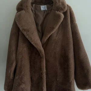 Brun pälsjacka med bred krage - Säljer en fluffig brun pälsjacka från NA-KD med bred krage och diskret knappstängning. Jackan har en mjuk, beige insida och är perfekt för dig som vill ha en cozy och trendig look under kyliga dagar. Passformen är loose och ärmarna är långa.