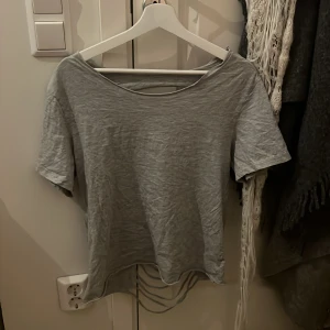 Grå t-shirt med öppen rygg - Säljer en grå t-shirt med korta ärmar och rund hals. Baksidan har en cool design med öppen rygg och snörning som ger en edgy vibe. Materialet känns mjukt och lätt, perfekt för dig som vill sticka ut med din stil. Har klippt den själv.
