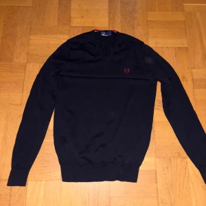 Svart v-ringad tröja från Fred Perry - Svart v-ringad tröja från Fred Perry med rött broderat logomärke på bröstet. Tröjan har långärmad passform och ribbade muddar vid ärmslut och nederkant. Perfekt för dig som gillar stilrena och klassiska plagg.