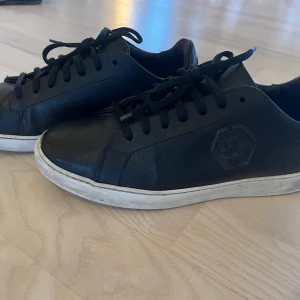 Svarta sneakers från Philipp Plein - Svarta sneakers i skinn från Philipp Plein med svart logga på sidan och klassisk vit platt sula. Skorna har snörning och en stilren, minimalistisk design som passar till det mesta. Perfekta för dig som gillar exklusiva och snygga sneakers. Låda och kvitto finns. Nypris 3519 kr.