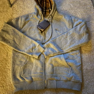 Grå hoodie från Burberry med dragkedja - Grå hoodie från Burberry med klassiskt rutigt foder i huvan och broderad logga på bröstet.storlek L men passar m