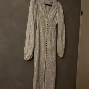 Adoore Positano Dress - Supersnygg vit långklänning från Adoore med blommigt broderat mönster, v-ringad hals och puffiga långärmar. Klänningen har knappar framtill och en slits som ger extra edge. Perfekt för dig som vill sticka ut med en romantisk och trendig look. Ord. pris 1695kr