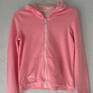 Rosa hoodie med dragkedja från H&M - Mysig ljusrosa hoodie från H&M Basic med dragkedja framtill och två fickor. Tröjan har huva, långa ärmar och ribbade muddar. Tillverkad i en mjuk blandning av bomull och polyester med lite stretch för extra komfort.