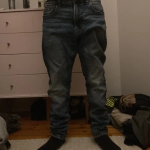 Blå tapered jeans - Säljer ett par blå jeans från New Yorker med tapered passform och klassisk femficksdesign. Jeansen har en mörk tvätt med snygga slitningar och är tillverkade i mjukt denimtyg. Perfekt för dig som gillar en avslappnad men ändå stilren look.