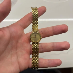 Guldfärgad Tissot klocka med länkarmband - Stilren guldfärgad klocka från Tissot med rund urtavla och klassiskt länkarmband i metall. Urtavlan är enkel och elegant utan siffror, och spännet har Tissots logga. Perfekt accessoar för dig som gillar tidlös design.