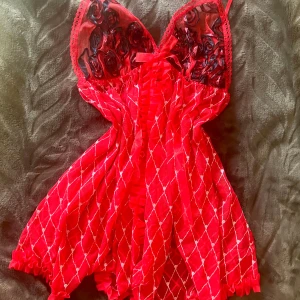 Röd topp med spets och rosett - Säljer en röd kortklänning med transparent mesh och vita rutmönster. Överdelen har svarta och röda broderade rosor, spetskant och en söt rosett framtill. Volangdetaljer längs mitten och nederkant ger extra flärd. Perfekt för dig som gillar unika detaljer.öppen i mitten men man kan fästa säkerhetsnål för att stänga 