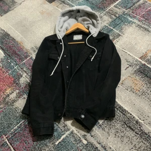 Svart jeansjacka med grå hoodie från SikSilk - Svart jeansjacka från SikSilk med fastsydd grå hoodie och vita snören. Jackan har klassiska jeansjacke-detaljer som bröstfickor med knappar och sidofickor. Snygg kontrast mellan svart denim och grått tyg i luvan, perfekt för en chill streetwear-look.