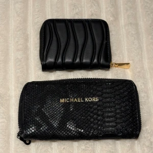 Svarta plånböcker, en Michael Kors - Två svarta plånböcker, en från Michael Kors med glansig ormskinnsstruktur och guldfärgad logga, samt en mindre plånbok i vågigt mönstrat skinn med guldfärgad dragkedja. Båda har dragkedja och stilren design, perfekta för kort och kontanter.
