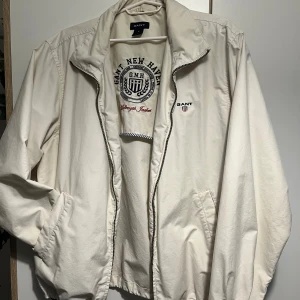Vindjacka från GANT - Snygg vit vindjacka från GANT med broderad logga på bröstet och coolt tryck på insidan. Passar perfekt till höst 🍂. Väldigt välvårdad men har använts ganska mycket.   OBS, Jag klippte mössan eftersom det var irriterande, men det kommer absolut inte förstöra utseendet av jackan tack vare plåstret som gömmer det (syns på bild 5!!).