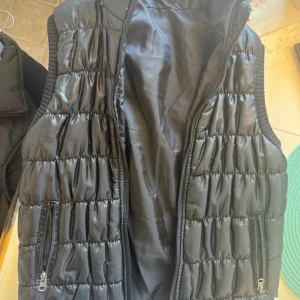 Svart quiltad dunväst med dragkedja - Snygg svart dunväst med quiltad design och glansig yta. Västen har två praktiska fickor med dragkedja och ribbade kanter vid ärmhålen. Perfekt att slänga över en hoodie för extra värme. Stängs med dragkedja framtill. Snabb leverans 📦 kolla gärna på mina andra plagg ! 