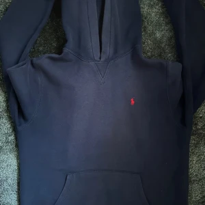 Mörkblå hoodie från Polo Ralph Lauren - Snygg mörkblå hoodie från Polo Ralph Lauren med klassisk röd logobrodyr på bröstet. Tröjan har huva, lång ärm och en stor magficka framtill. Perfekt för dig som gillar stilren design och vill ha en hoodie med premiumkänsla.