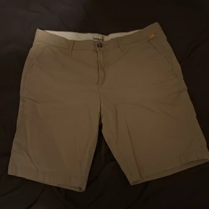 Beige shorts från Timberland W36 - Snygga beige shorts från Timberland i klassisk modell med raka ben och fickor både fram och bak. Sköna och stilrena, perfekta för sommardagar. Tillverkade i mjuk bomull och har bälteshällor samt knapp och dragkedja framtill.