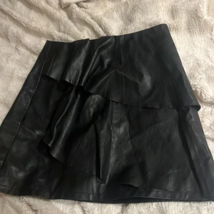 Svart skinnkjol med volang - Säljer en svart kort skinnkjol med snygga volangdetaljer framtill. Kjolen har en cool och edgy vibe, perfekt för att lyfta din outfit. Materialet är mjukt och följsamt, och volangen ger kjolen extra rörelse och stil.