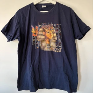 Mörkblå King Tut Egypt t-shirt - Mörkblå t-shirt med färgglatt tryck av farao Tutankhamon och egyptiska hieroglyfer på bröstet. Klassisk passform med rund hals och korta ärmar. Tillverkad i 100% bomull, skön och mjuk mot huden. Perfekt för dig som gillar unika motiv och historia. Själva trycket är lite sådär sprucket 