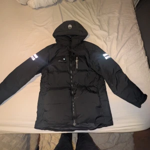 Svart Sail Racing pufferjacka  - Svart pufferjacka från Sail Racing, modell Antarctica Expedition. Jackan har huva, reflekterande detaljer på ärmarna och logga på bröstet. Utrustad med dragkedja framtill och flera praktiska fickor. Perfekt för kalla dagar och har en clean, sportig look.