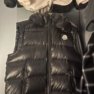 Svart dunväst från Moncler med huva - Svart dunväst från Moncler med avtagbar huva och två dragkedjeförsedda fickor framtill. Västen har quiltad design och Moncler-logga på bröstet. Insidan har en unik serietidningsetikett. Perfekt för att hålla värmen och se stilren ut.